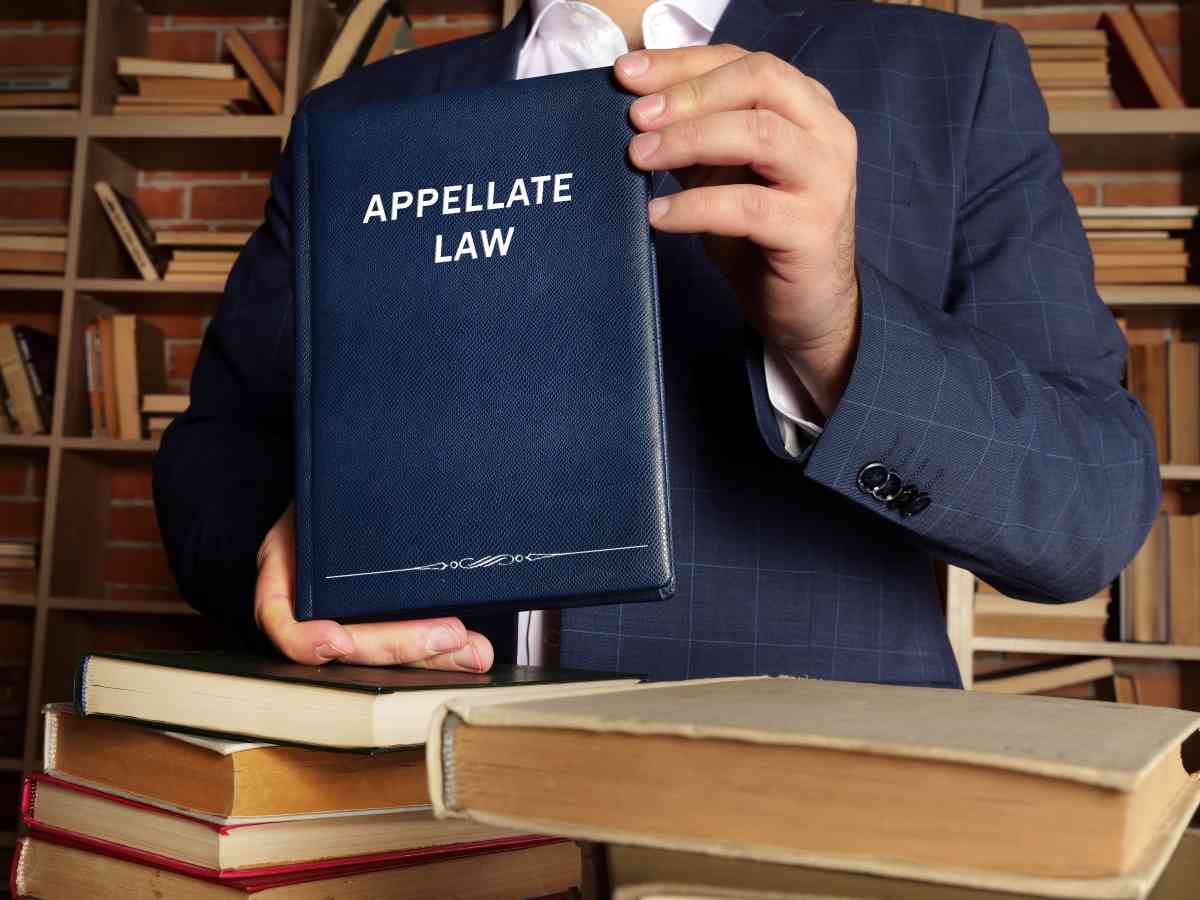 A person in a navy blue suit holds up a dark blue book titled 'Appellate Law' in a library setting, with stacks of books in the foreground and bookshelves in the background.