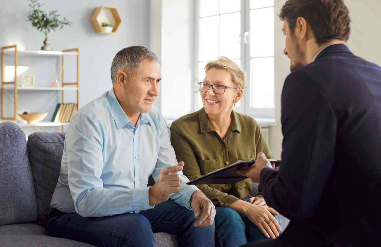 A middle-aged couple sitting on a sofa in conversation with a professional holding a clipboard, during a legal consultation meeting.